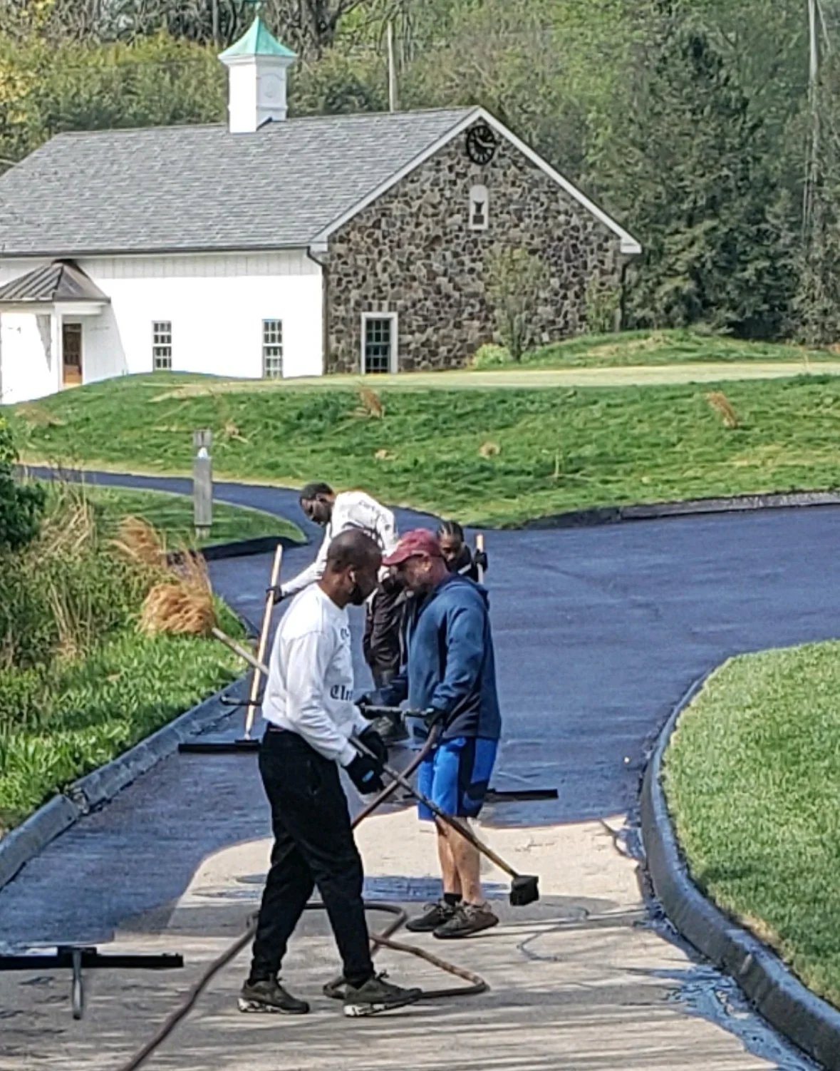 Crew applying dark blue sealcoat to residential driveway with stone farmhouse in background